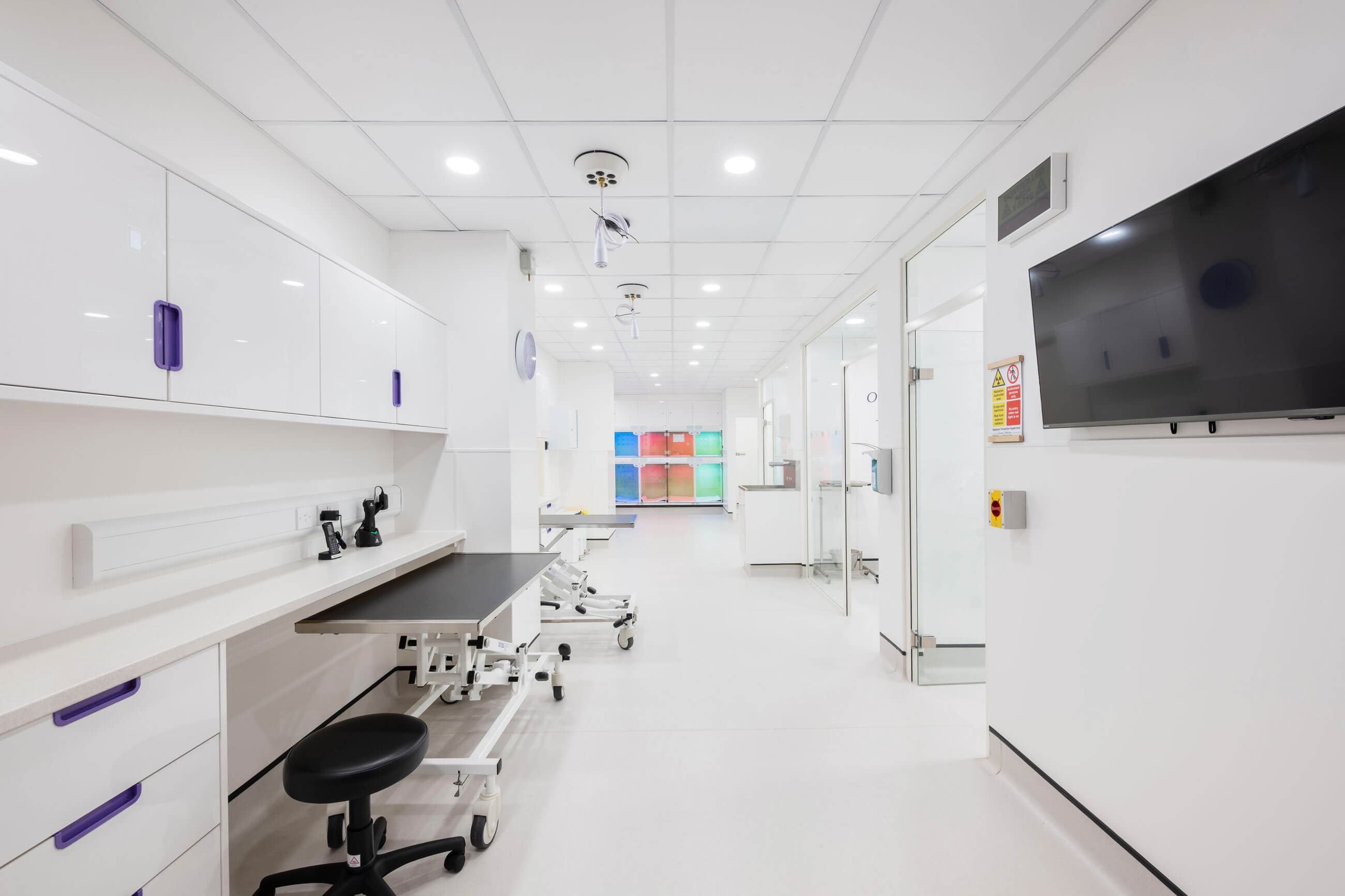 Modern, sterile medical room with white cabinetry, adjustable furniture, and colorful equipment storage in the background. Brightly lit environment.