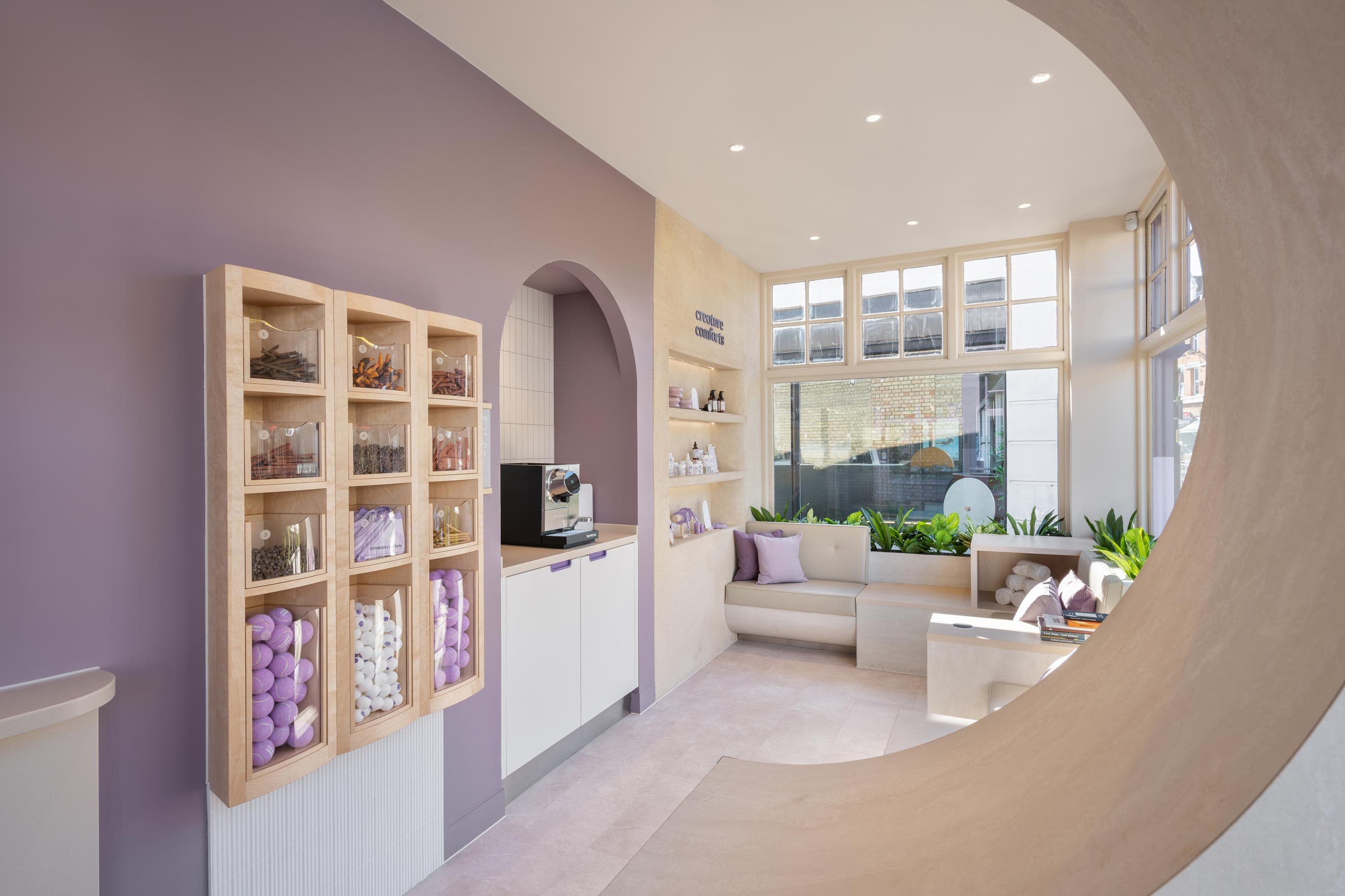 Bright indoor space featuring a lavender wall, wooden display of colorful sweets, coffee station, and cozy seating area with plants.